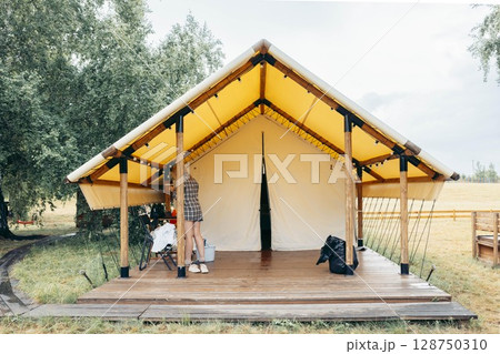 woman standing in front of modern glamping tent adjusting entrance cover at wooden platform, trees, dry grass in background, tourism, sustainable travel, outdoor lifestyle 128750310