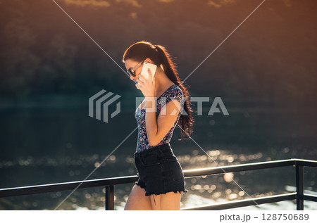 The smiling girl in shorts and a tank top is happily talking on the phone with the shimmering water in the background The smiling girl in shorts and a tank top is happily talking on the phone with the shimmering water in the background 128750689