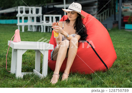 A young girl with a hat is sitting comfortably in a red beanbag chair on the grass A young girl with a hat is sitting comfortably in a red beanbag chair on the grass 128750789