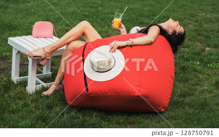A stylish girl in a hat is sitting on a red beanbag chair and slowly drinking juice 128750791
