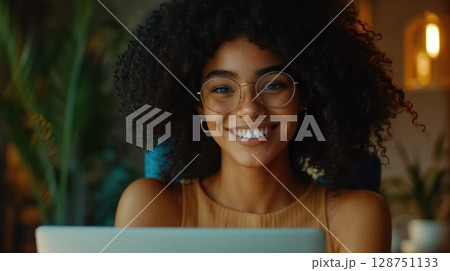 Young woman with natural afro hair wearing round glasses smiles warmly at laptop in cozy home environment. Soft ambient lighting creates intimate atmosphere while houseplants add organic backdrop 128751133