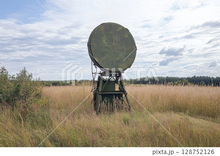 Large military radar dish in grassy field, surrounded by tall grass and trees, cloudy sky overhead, communication technology 128752126