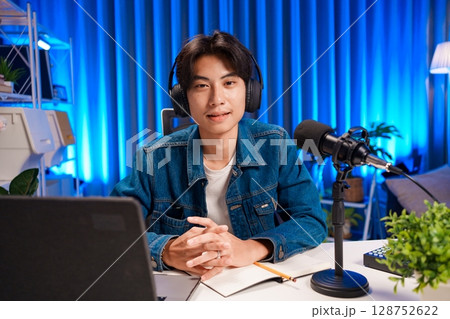 Man podcaster influencer blogger smiling while broadcasting his live audio podcast in studio using headphones. Male radio host making podcast or interview. 128752622