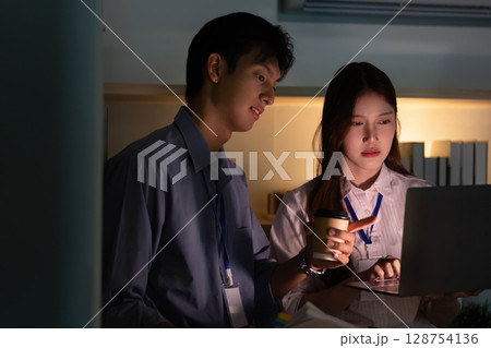 Teamwork and Technology. Colleagues collaborating on a laptop in a dimly lit office. Teamwork and Technology. Colleagues collaborating on a laptop in a dimly lit office. 128754136