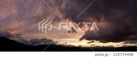 Dramatic sunset scene with dark, brooding clouds casting shadows over sky. Horizon glows with warm hues of orange and pink, contrasting against silhouettes of mountains, creating moody atmosphere. 128754406