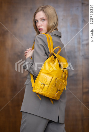 A stylish young woman, long blonde hair, poses in a grey suit, holding a vibrant yellow leather backpack against a warm wooden background 128756314