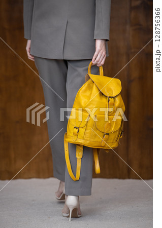 A stylish young woman, poses in a grey suit, holding a vibrant yellow leather backpack against a warm wooden background A stylish young woman, poses in a grey suit, holding a vibrant yellow leather backpack against a warm wooden background 128756366