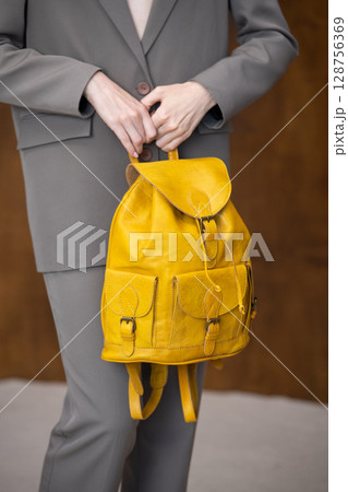 A stylish young woman, poses in a grey suit, holding a vibrant yellow leather backpack against a warm wooden background 128756369