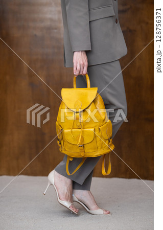 A stylish young woman, poses in a grey suit, holding a vibrant yellow leather backpack against a warm wooden background 128756371