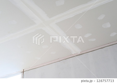 Interior building renovation progress showing new gypsum board ceiling with smooth edges ready for plastering. Electrical wire visible before final smooth finish applied to ceiling surface 128757713