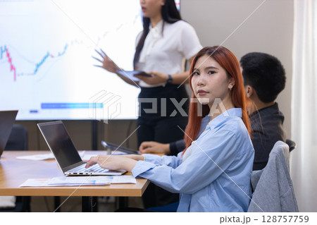 Women with red hair attentively works on her laptop while colleague delivers presentation. Women with red hair attentively works on her laptop while colleague delivers presentation. 128757759