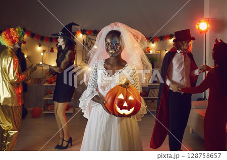 Happy black woman dressed as Dead Bride with her jack-o-lantern at Halloween party 128758657