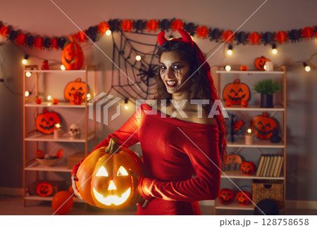 Beautiful woman who's dressed as devil holding Halloween jack-o-lantern and smiling 128758658