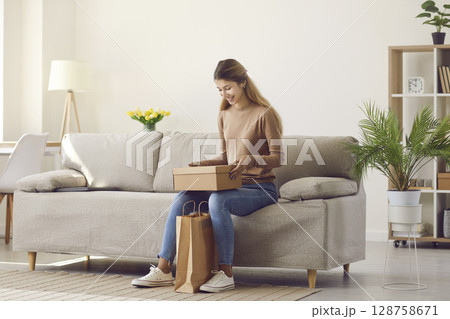 Happy young woman sitting on sofa at home unpacks cardboard parcel with online order from internet. 128758671