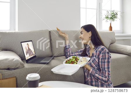 Happy woman talking to friend on video call while eating takeaway meal on lunch break Happy woman talking to friend on video call while eating takeaway meal on lunch break 128758673