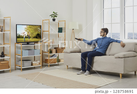 Young man relaxing on sofa in living room and watching film on large screen of modern TV 128758691