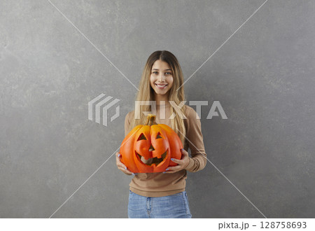 Studio portrait of young woman holding pumpkin with carved face in style of Halloween Studio portrait of young woman holding pumpkin with carved face in style of Halloween 128758693