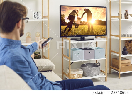 Relaxed man sitting on a couch at home and watching a movie on a large TV screen Relaxed man sitting on a couch at home and watching a movie on a large TV screen 128758695
