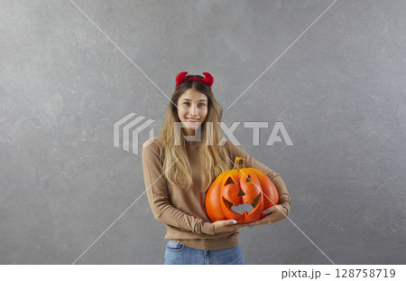 Portrait of happy young woman or teenage girl in devil horns holding Halloween pumpkin 128758719