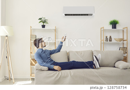 Man sitting on couch at home and using remote control to turn on air conditioner Man sitting on couch at home and using remote control to turn on air conditioner 128758734