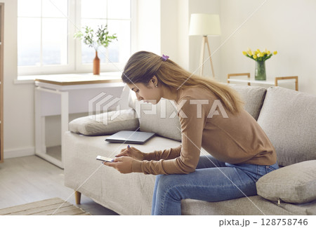 Woman sitting on sofa, holding cellphone, browsing Internet or chatting on social media Woman sitting on sofa, holding cellphone, browsing Internet or chatting on social media 128758746