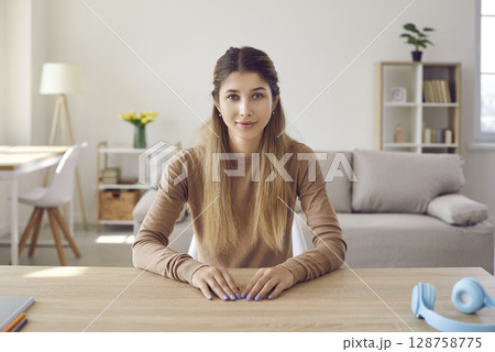 Portrait of cute friendly young woman who is looking at webcam while sitting at home in living room. 128758775