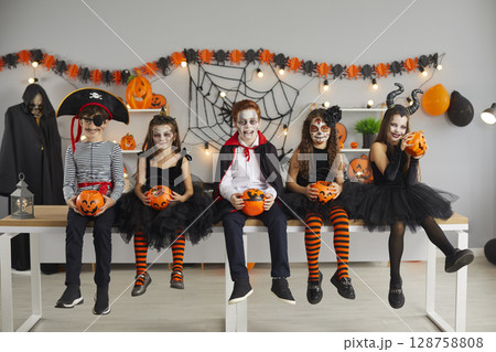 Children dressed in various spooky characters sit in row holding pumpkin buckets for candy. Children dressed in various spooky characters sit in row holding pumpkin buckets for candy. 128758808