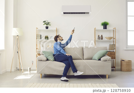Man who's sitting on couch at home switches air conditioner using remote control Man who's sitting on couch at home switches air conditioner using remote control 128758910