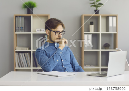 Portrait of concentrated young man thinking while sitting in office in front of laptop. Portrait of concentrated young man thinking while sitting in office in front of laptop. 128758992