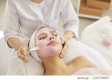 Young woman or teen girl enjoying cosmetic clay facial mask in spa and beauty salon 128759007