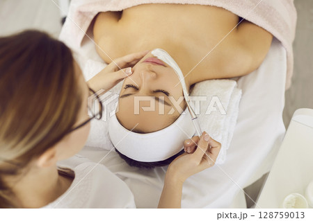 Cosmetologist at beauty salon applying cosmetic clay mask on face of young woman Cosmetologist at beauty salon applying cosmetic clay mask on face of young woman 128759013