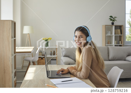 Happy teen girl in headphones at home preparing homework sitting with notebook at laptop. Happy teen girl in headphones at home preparing homework sitting with notebook at laptop. 128759216