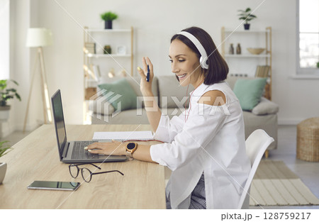 Woman in headphones waving hello at the laptop screen at the start of a video call Woman in headphones waving hello at the laptop screen at the start of a video call 128759217