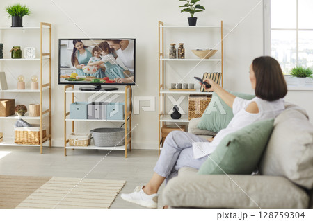 Woman sitting on sofa at home and switching on her television set with big screen 128759304
