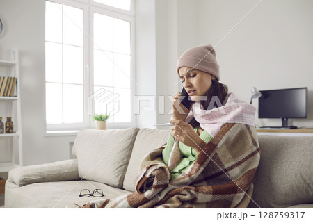 Woman who has flu or cold sitting on sofa, looking at thermometer and calling doctor 128759317