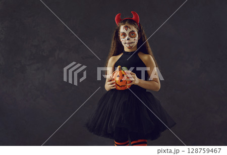 Studio portrait of a little girl with a skull makeup holding a Halloween pumpkin 128759467