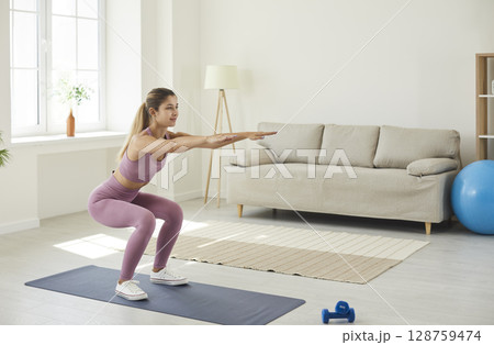 Young enduring woman trains at home doing deep squats with her arms outstretched. Young enduring woman trains at home doing deep squats with her arms outstretched. 128759474