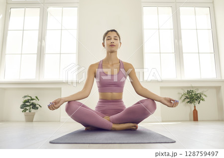 Calm relaxed young lady sitting on yoga mat in gym and doing meditation exercise Calm relaxed young lady sitting on yoga mat in gym and doing meditation exercise 128759497