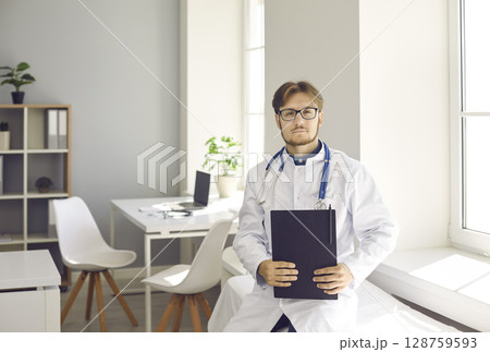 Portrait of confident young male doctor standing in office of modern medical center. Portrait of confident young male doctor standing in office of modern medical center. 128759593