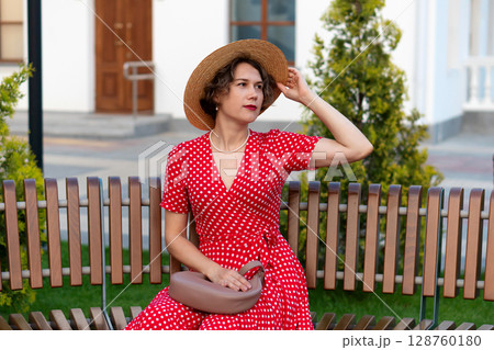 Woman in red polka-dot dress adjusts hat while seated outdoors. Woman in red polka-dot dress adjusts hat while seated outdoors. 128760180