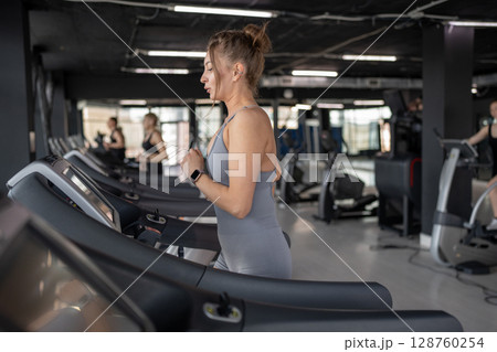 A young woman runs on a treadmill in a gym, showing her dedication to fitness and wellbeing 128760254