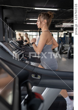 A young woman runs on a treadmill in a gym, showing her dedication to fitness and wellbeing A young woman runs on a treadmill in a gym, showing her dedication to fitness and wellbeing 128760316
