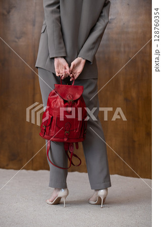 A stylish young woman, poses in a grey suit, holding a vibrant red leather backpack against a warm wooden background 128760354