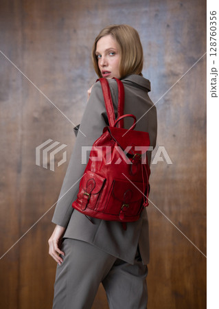 A stylish young woman, long blonde hair, poses in a grey suit, holding a vibrant red leather backpack against a warm wooden background A stylish young woman, long blonde hair, poses in a grey suit, holding a vibrant red leather backpack against a warm wooden background 128760356