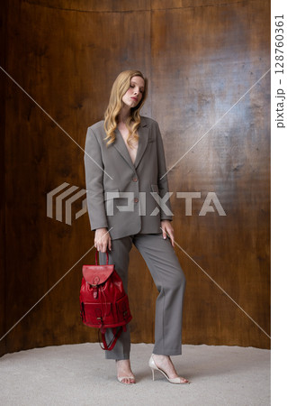 A stylish young woman, long blonde hair, poses in a grey suit, holding a vibrant red leather backpack against a warm wooden background A stylish young woman, long blonde hair, poses in a grey suit, holding a vibrant red leather backpack against a warm wooden background 128760361
