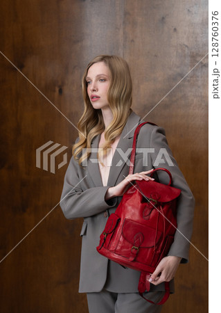 A stylish young woman, long blonde hair, poses in a grey suit, holding a vibrant red leather backpack against a warm wooden background A stylish young woman, long blonde hair, poses in a grey suit, holding a vibrant red leather backpack against a warm wooden background 128760376