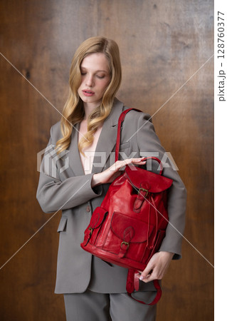 A stylish young woman, long blonde hair, poses in a grey suit, holding a vibrant red leather backpack against a warm wooden background 128760377