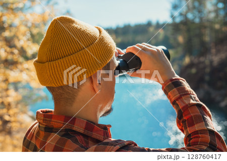 Man ecologist wearing a yellow beanie and plaid shirt is observing nature through binoculars, standing by serene lake surrounded by trees, outdoor exploration, birdwatching 128760417