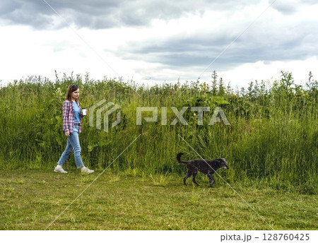 Woman with cup of coffee in hand walking mixed breed gray fluffy senior dog bedlington terrier whippet through grassy field with tall green grass and cloudy sky in the background 128760425