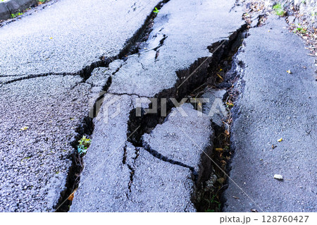Cracked asphalt road surface damaged by landslide, showcasing deep fissures and uneven terrain, illustrating the impact of natural disasters on infrastructure and environment Cracked asphalt road surface damaged by landslide, showcasing deep fissures and uneven terrain, illustrating the impact of natural disasters on infrastructure and environment 128760427